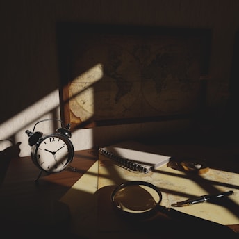 A dimly lit desk with an antique alarm clock showing seven o'clock, a spiral notebook, a magnifying glass, and a pen. A vintage world map on the wall casts a shadow over the workspace, creating a mysterious and contemplative atmosphere.