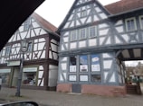 Two traditional half-timbered buildings with intricate wooden frameworks and white plaster walls are situated next to each other. The structures feature gabled roofs covered with reddish-brown tiles. A storefront sign indicates a medical and orthopedic practice inside one of the buildings. A classic street lamp is visible on the cobblestone sidewalk in front of the buildings.