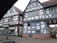 Two traditional half-timbered buildings with intricate wooden frameworks and white plaster walls are situated next to each other. The structures feature gabled roofs covered with reddish-brown tiles. A storefront sign indicates a medical and orthopedic practice inside one of the buildings. A classic street lamp is visible on the cobblestone sidewalk in front of the buildings.