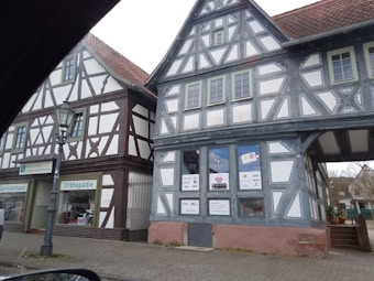 Two traditional half-timbered buildings with intricate wooden frameworks and white plaster walls are situated next to each other. The structures feature gabled roofs covered with reddish-brown tiles. A storefront sign indicates a medical and orthopedic practice inside one of the buildings. A classic street lamp is visible on the cobblestone sidewalk in front of the buildings.