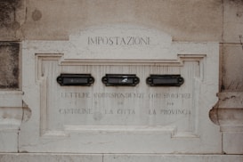 An old stone postal box embedded in a wall with three slots labeled for different types of correspondence. The text is in Italian, with inscriptions above each slot for specific categories.
