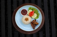 A plate of rice garnished with fried shallots accompanied by grilled chicken wings, sliced cucumber, tomato, and a leaf of lettuce. A small bowl of spicy sambal sauce is placed on the side. The setting is on a wooden table with a blue plate.
