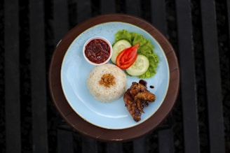A plate of rice garnished with fried shallots accompanied by grilled chicken wings, sliced cucumber, tomato, and a leaf of lettuce. A small bowl of spicy sambal sauce is placed on the side. The setting is on a wooden table with a blue plate.