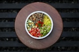 A round foil container filled with a colorful arrangement of ingredients including diced cucumbers, tomatoes, lettuce, yellow rice, and a brown grain mixture. The container is placed on a wooden surface with slatted patterns visible in the background.