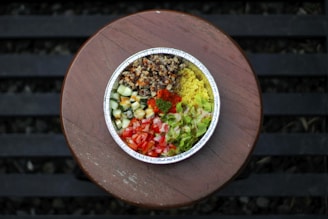 A round foil container filled with a colorful arrangement of ingredients including diced cucumbers, tomatoes, lettuce, yellow rice, and a brown grain mixture. The container is placed on a wooden surface with slatted patterns visible in the background.