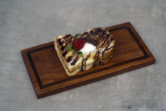 A wooden tray holds a small stack of toasted bread slices drizzled with chocolate sauce and garnished with a strawberry, grated white cheese, and mint leaves.