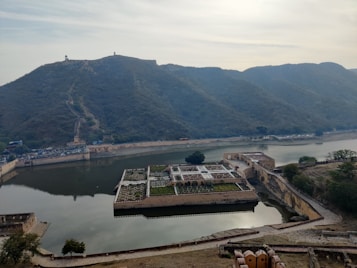A large, historic fort sits atop a hill, surrounded by lush greenery and overlooking a body of water. The landscape includes a series of terraces and stone structures that extend into the water. The fort features intricate pathways and gardens, creating an impressive architectural site. The sky is overcast, adding a serene quality to the environment.