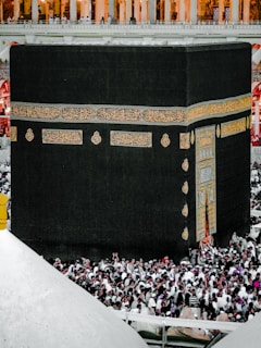 A large crowd of people surrounding the Kaaba, a black cubic structure adorned with Arabic calligraphy in gold. The scene is set in a mosque environment with an upper balcony visible.