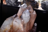 A cluster of large, clear crystal formations with distinct pointed tips is prominently displayed. The crystals exhibit a range of natural hues from transparent to light brown, with some earthy inclusions visible within. The lighting emphasizes the facets and angles of the crystals, creating reflections and subtle color variations.