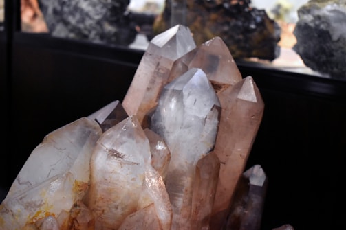 A cluster of large, clear crystal formations with distinct pointed tips is prominently displayed. The crystals exhibit a range of natural hues from transparent to light brown, with some earthy inclusions visible within. The lighting emphasizes the facets and angles of the crystals, creating reflections and subtle color variations.
