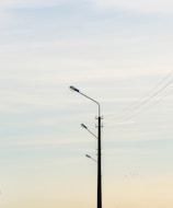 A tall street lamp post with three lamps aligned vertically. Electric wires are extending from the pole, set against a soft gradient sky transitioning from light blue to pale yellow. A small flock of birds is visible in the distance, adding a serene element to the scene.