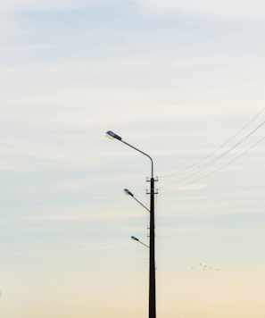 A tall street lamp post with three lamps aligned vertically. Electric wires are extending from the pole, set against a soft gradient sky transitioning from light blue to pale yellow. A small flock of birds is visible in the distance, adding a serene element to the scene.