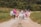 A group of seven people are walking together along a gravel path in a rural setting. They appear to be friends, with some linking arms and others holding hands. The group is casually dressed in summer clothing, and they seem happy and relaxed. The path is flanked by green grass and trees are visible in the background.