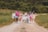 A group of seven people are walking together along a gravel path in a rural setting. They appear to be friends, with some linking arms and others holding hands. The group is casually dressed in summer clothing, and they seem happy and relaxed. The path is flanked by green grass and trees are visible in the background.