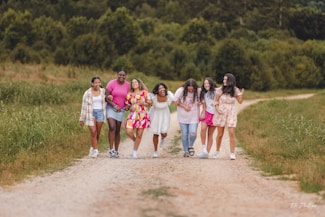A group of seven people are walking together along a gravel path in a rural setting. They appear to be friends, with some linking arms and others holding hands. The group is casually dressed in summer clothing, and they seem happy and relaxed. The path is flanked by green grass and trees are visible in the background.