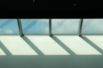 Sunlight streams through a row of skylight windows, casting geometric shadows on a white ceiling. The sky outside is partially visible, showing a light blue hue with scattered clouds.