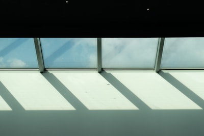Sunlight streams through a row of skylight windows, casting geometric shadows on a white ceiling. The sky outside is partially visible, showing a light blue hue with scattered clouds.