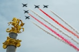 A group of military jets fly in formation, leaving trails of red and white smoke across a clear sky. In the foreground, a golden statue of a horse and rider is prominently displayed atop a pedestal.