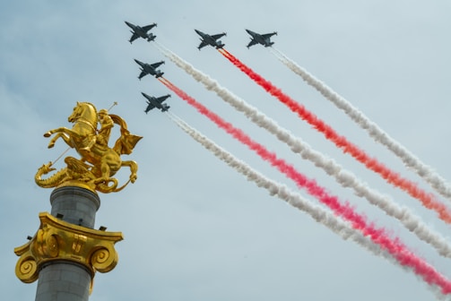 A group of military jets fly in formation, leaving trails of red and white smoke across a clear sky. In the foreground, a golden statue of a horse and rider is prominently displayed atop a pedestal.