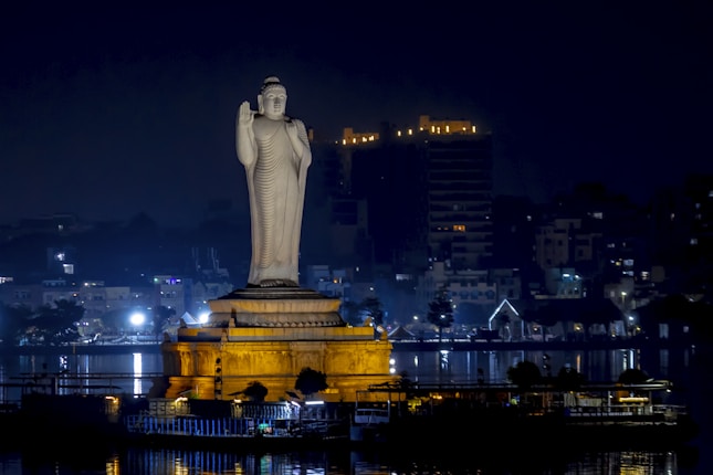 A large Buddha statue stands majestically on a pedestal, illuminated against the night sky. The backdrop features a cityscape with dimly lit buildings and street lights reflecting off a body of water.