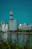 A cityscape featuring a prominent red and white communication tower amidst several modern buildings. In the foreground, there is a body of water reflecting the structures and a few trees. The sky is clear and blue, providing a vibrant backdrop.