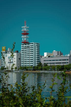 A cityscape featuring a prominent red and white communication tower amidst several modern buildings. In the foreground, there is a body of water reflecting the structures and a few trees. The sky is clear and blue, providing a vibrant backdrop.