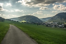 A picturesque landscape with a narrow road leading through expansive green fields toward a small village nestled between lush, forested hills. The sky is partly cloudy, and the sunlight filters through the clouds, casting soft shadows on the ground.