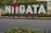 Large white letters spelling out 'NIIGATA' are displayed outdoors with two heart symbols above the 'I's. The letters are placed against a backdrop of green shrubbery, with surrounding urban elements like a road and buildings.