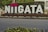 Large white letters spelling out 'NIIGATA' are displayed outdoors with two heart symbols above the 'I's. The letters are placed against a backdrop of green shrubbery, with surrounding urban elements like a road and buildings.