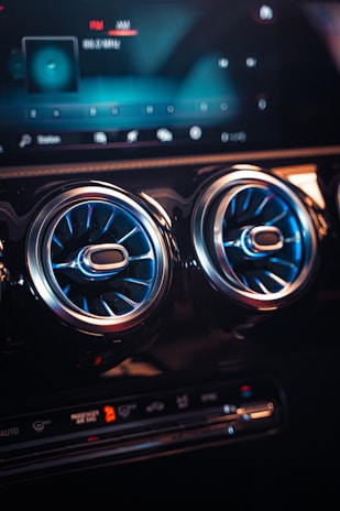 The image depicts the interior of a modern car focusing on the dashboard and air vents. The dashboard display screen shows various digital interfaces, including a radio display, with a blue and black color scheme. Two circular air vents with a metallic and blue design are prominently visible, and the lower dashboard controls include climate control buttons and displays, with a mix of illuminated and dark features.