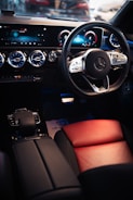 The interior of a luxury car is highlighted, featuring a sleek steering wheel with a premium brand logo, a modern digital dashboard display with blue accents, and ventilated air conditioning vents. The seating includes a combination of black and red leather, contributing to a sophisticated and elegant design.