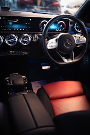 The interior of a luxury car is highlighted, featuring a sleek steering wheel with a premium brand logo, a modern digital dashboard display with blue accents, and ventilated air conditioning vents. The seating includes a combination of black and red leather, contributing to a sophisticated and elegant design.