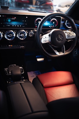 The interior of a luxury car is highlighted, featuring a sleek steering wheel with a premium brand logo, a modern digital dashboard display with blue accents, and ventilated air conditioning vents. The seating includes a combination of black and red leather, contributing to a sophisticated and elegant design.