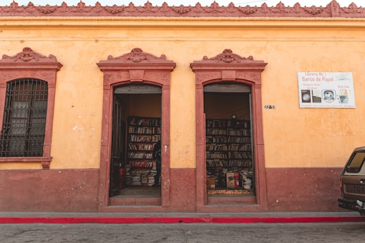 A small bookstore with two open doorways leading to shelves filled with books. The building has a rustic yellow facade with reddish-brown trim and architectural details. A sign next to the doorway reads 'Librería No 1 Barco de Papel' advertising book sales.