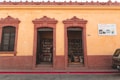 A small bookstore with two open doorways leading to shelves filled with books. The building has a rustic yellow facade with reddish-brown trim and architectural details. A sign next to the doorway reads 'Librería No 1 Barco de Papel' advertising book sales.