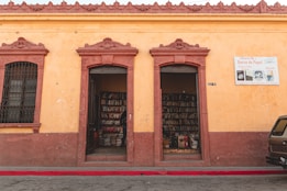 A small bookstore with two open doorways leading to shelves filled with books. The building has a rustic yellow facade with reddish-brown trim and architectural details. A sign next to the doorway reads 'Librer&iacute;a No 1 Barco de Papel' advertising book sales.