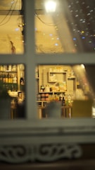 A view through a window showcasing shelves filled with various food and drink products. The setting appears cozy with warm lighting and decorative elements, including a white basket and some bottles. The window frame divides the scene, creating a sense of looking into a quaint shop or cafe.