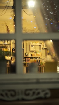 A view through a window showcasing shelves filled with various food and drink products. The setting appears cozy with warm lighting and decorative elements, including a white basket and some bottles. The window frame divides the scene, creating a sense of looking into a quaint shop or cafe.