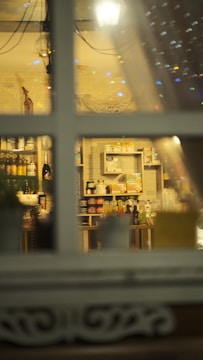 A view through a window showcasing shelves filled with various food and drink products. The setting appears cozy with warm lighting and decorative elements, including a white basket and some bottles. The window frame divides the scene, creating a sense of looking into a quaint shop or cafe.