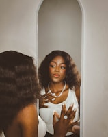 A person with long, wavy hair gazes into a mirror with an expression of contemplation. They are wearing a white top and a sparkling necklace, and have intricately designed nails adorned with rings. The setting appears serene with a soft color palette.