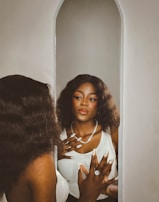 A person with long, wavy hair gazes into a mirror with an expression of contemplation. They are wearing a white top and a sparkling necklace, and have intricately designed nails adorned with rings. The setting appears serene with a soft color palette.