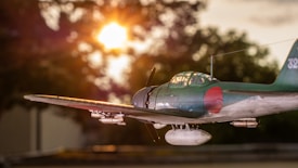 A vintage fighter aircraft is captured in flight against the backdrop of a vibrant sunset. The sun is low in the sky, casting a golden glow that highlights the details on the aircraft. The plane’s design includes a red circular emblem on its side and wing. Silhouetted trees are visible in the background, adding depth to the scene.