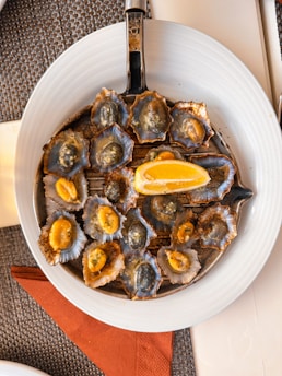 A white plate contains a round metal grill filled with small shellfish cooked in their shells, topped with a piece of lemon. The shells have a mix of dark blue and gray hues with bright orange flesh visible in some. A woven mat and brown napkin are visible underneath the plate.