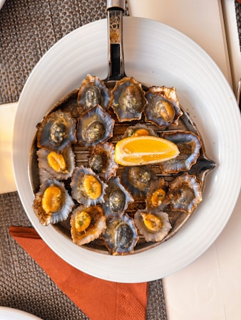 A white plate contains a round metal grill filled with small shellfish cooked in their shells, topped with a piece of lemon. The shells have a mix of dark blue and gray hues with bright orange flesh visible in some. A woven mat and brown napkin are visible underneath the plate.