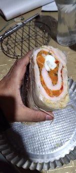 A hand is holding a sliced roll that consists of layers of meat, orange filling, and a white center. The roll is placed on a ridged metal tray with a clear plastic sheet partially covering it. In the background, there is a glass with water and kitchen utensils.