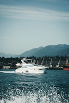 A luxury white yacht glides smoothly over the water with waves splashing in the foreground. The backdrop features a range of green hills under a partly cloudy sky. Several cranes are visible on the dockside, indicating an industrial area along the shoreline.