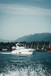 A luxury white yacht glides smoothly over the water with waves splashing in the foreground. The backdrop features a range of green hills under a partly cloudy sky. Several cranes are visible on the dockside, indicating an industrial area along the shoreline.