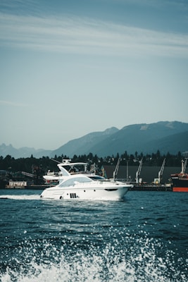 A luxury white yacht glides smoothly over the water with waves splashing in the foreground. The backdrop features a range of green hills under a partly cloudy sky. Several cranes are visible on the dockside, indicating an industrial area along the shoreline.