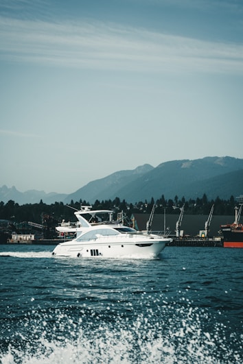 A luxury white yacht glides smoothly over the water with waves splashing in the foreground. The backdrop features a range of green hills under a partly cloudy sky. Several cranes are visible on the dockside, indicating an industrial area along the shoreline.