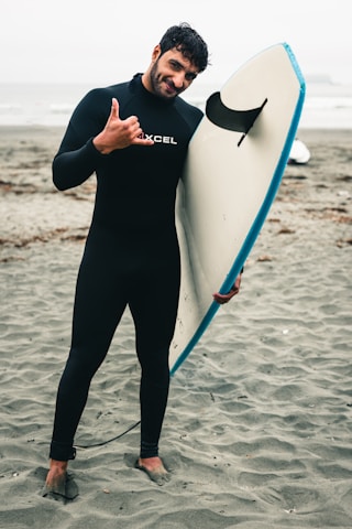 A person in a black wetsuit stands on a sandy beach, holding a surfboard upright. The surfboard has a blue edge and is resting against the individual's side. The person is making a shaka sign with their right hand, smiling slightly, and their feet are partially buried in the sand. The ocean and a cloudy sky are visible in the background.